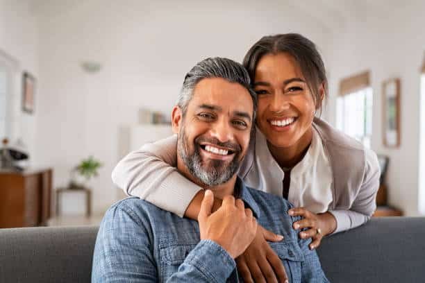 Smiling man sitting on a couch with a woman standing behind him, arms around his shoulders; both are looking at the camera in a bright living room.