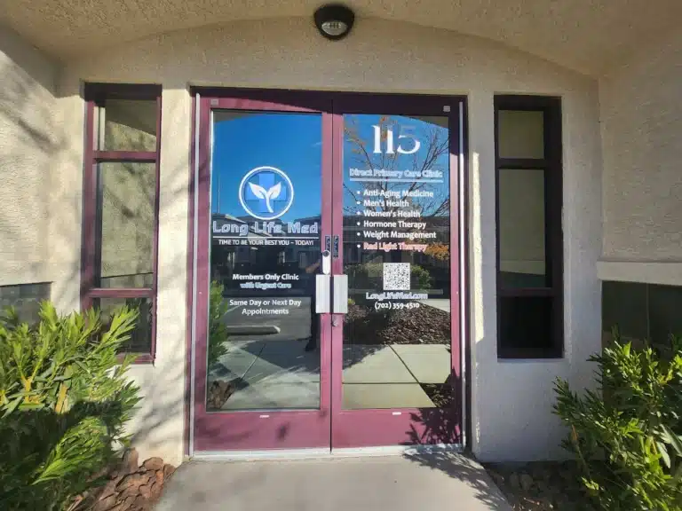 Glass doors of a Las Vegas medical clinic with "Long Life Med" logo, urgent care services list, clinic hours, website, phone number, and office number 115, surrounded by plants and sunlight.