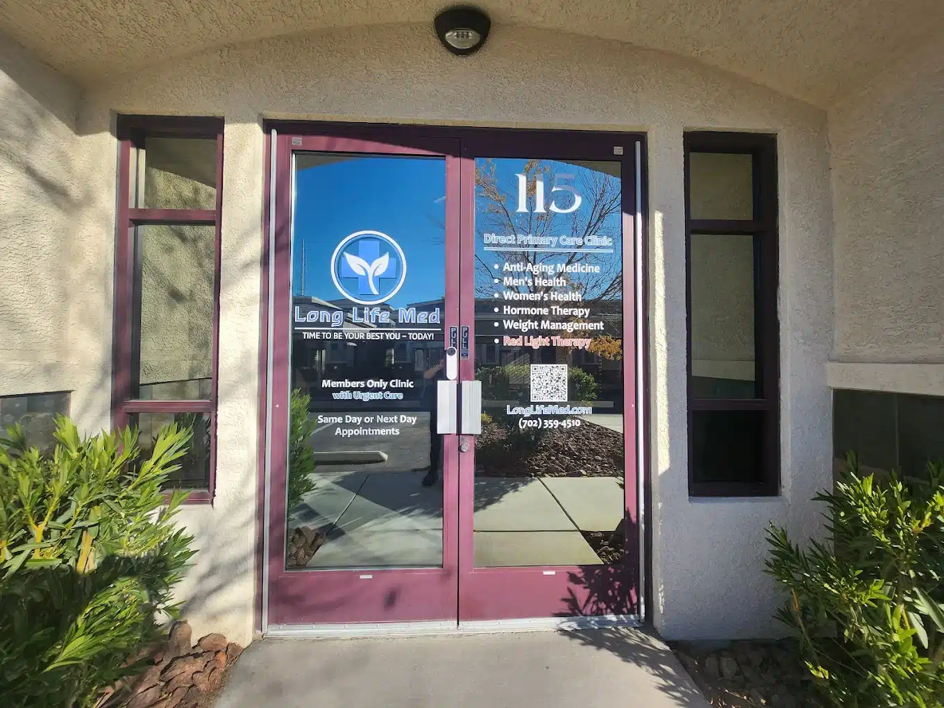 Glass doors of a Las Vegas medical clinic with "Long Life Med" logo, urgent care services list, clinic hours, website, phone number, and office number 115, surrounded by plants and sunlight.