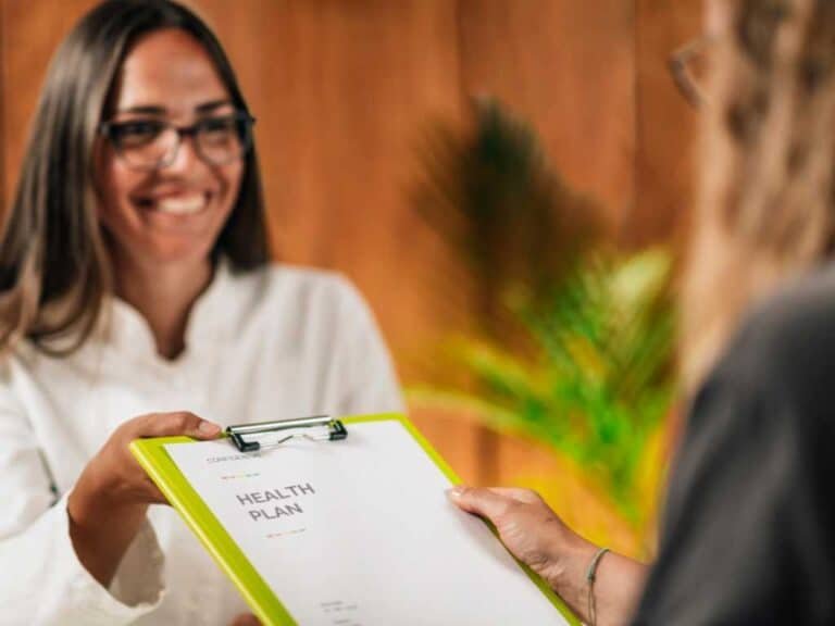 A woman hands a clipboard labeled "Health Plan" to another person in an indoor setting, helping them find a functional medicine doctor in Las Vegas.