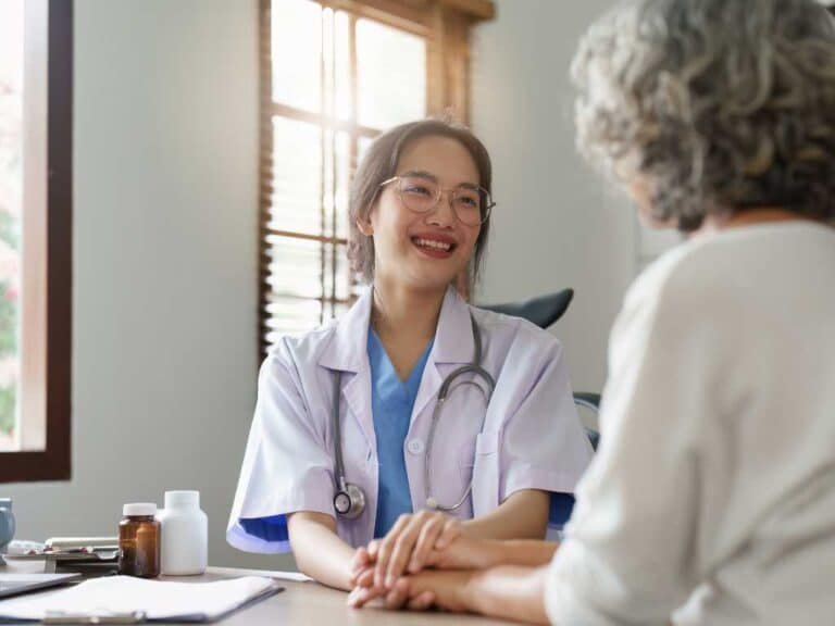 A female doctor at a Las Vegas Direct Primary Care clinic smiles and holds hands with an older patient during a consultation in a medical office.