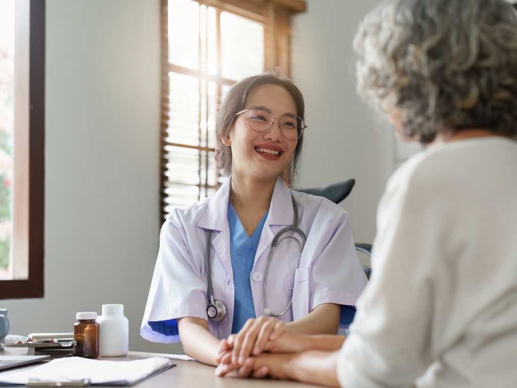 A female doctor at a Las Vegas Direct Primary Care clinic smiles and holds hands with an older patient during a consultation in a medical office.
