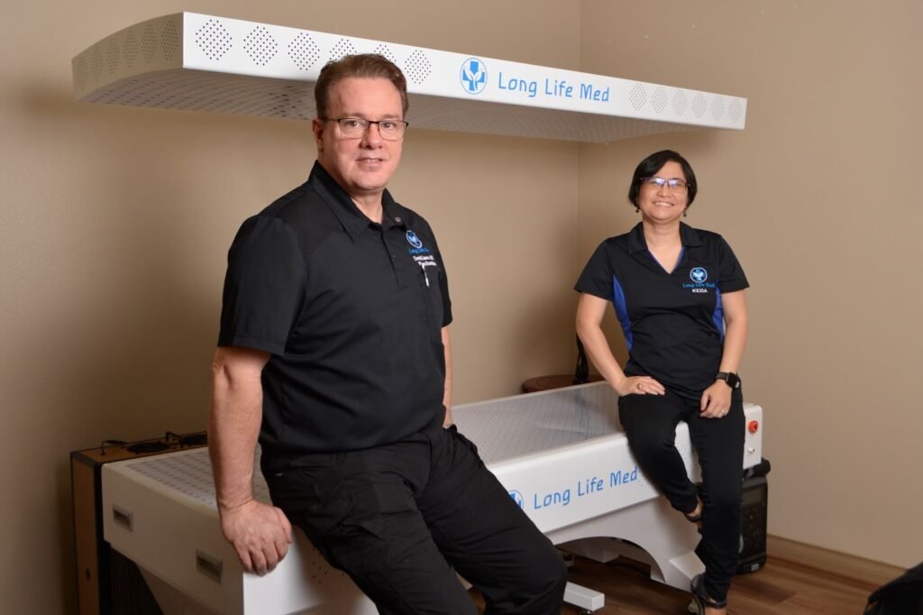Two people wearing black uniforms with "Long Life Med" logos sit and stand near a white medical device labeled "Long Life Med" in an indoor room with beige walls.