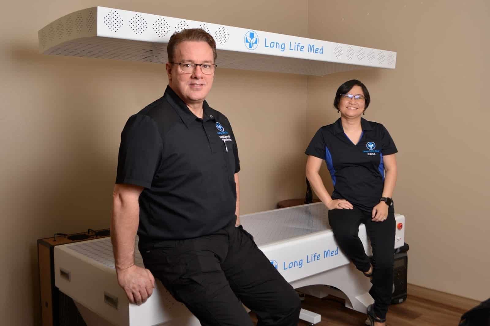 Two people wearing black uniforms with "Long Life Med" logos sit and stand near a white medical device labeled "Long Life Med" in an indoor room with beige walls.