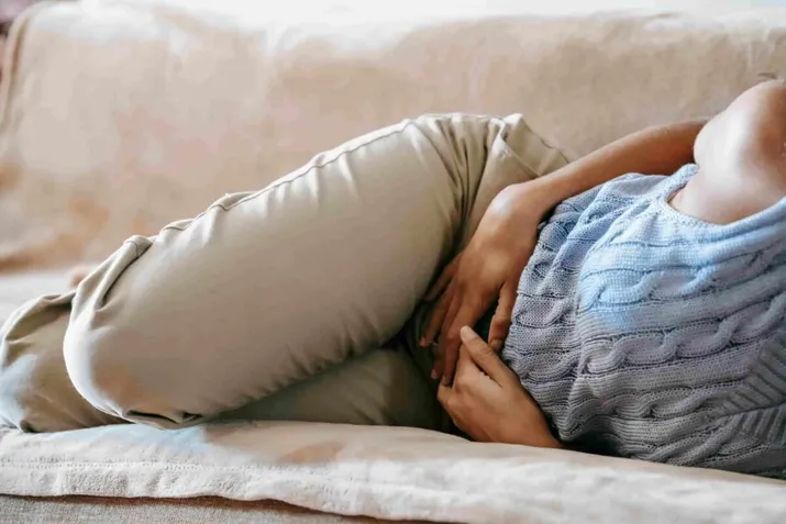 A person lying on a couch, wearing beige pants and a blue sweater, holding their stomach with both hands—possibly seeking Functional Medicine for Gastro Disease relief.