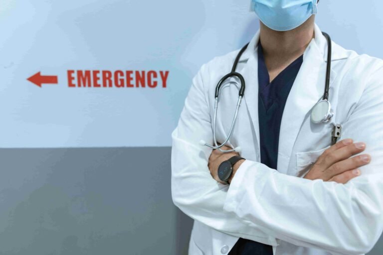 A healthcare professional in a lab coat and stethoscope stands with arms crossed near a sign pointing to the emergency department, emphasizing access to urgent care without insurance.