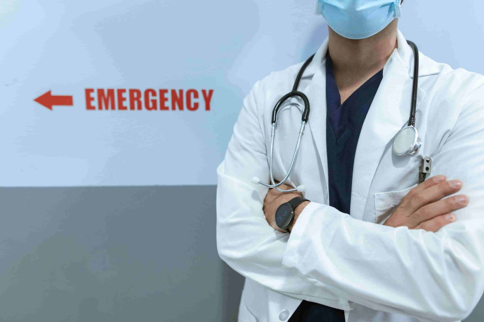 A healthcare professional in a lab coat and stethoscope stands with arms crossed near a sign pointing to the emergency department, emphasizing access to urgent care without insurance.