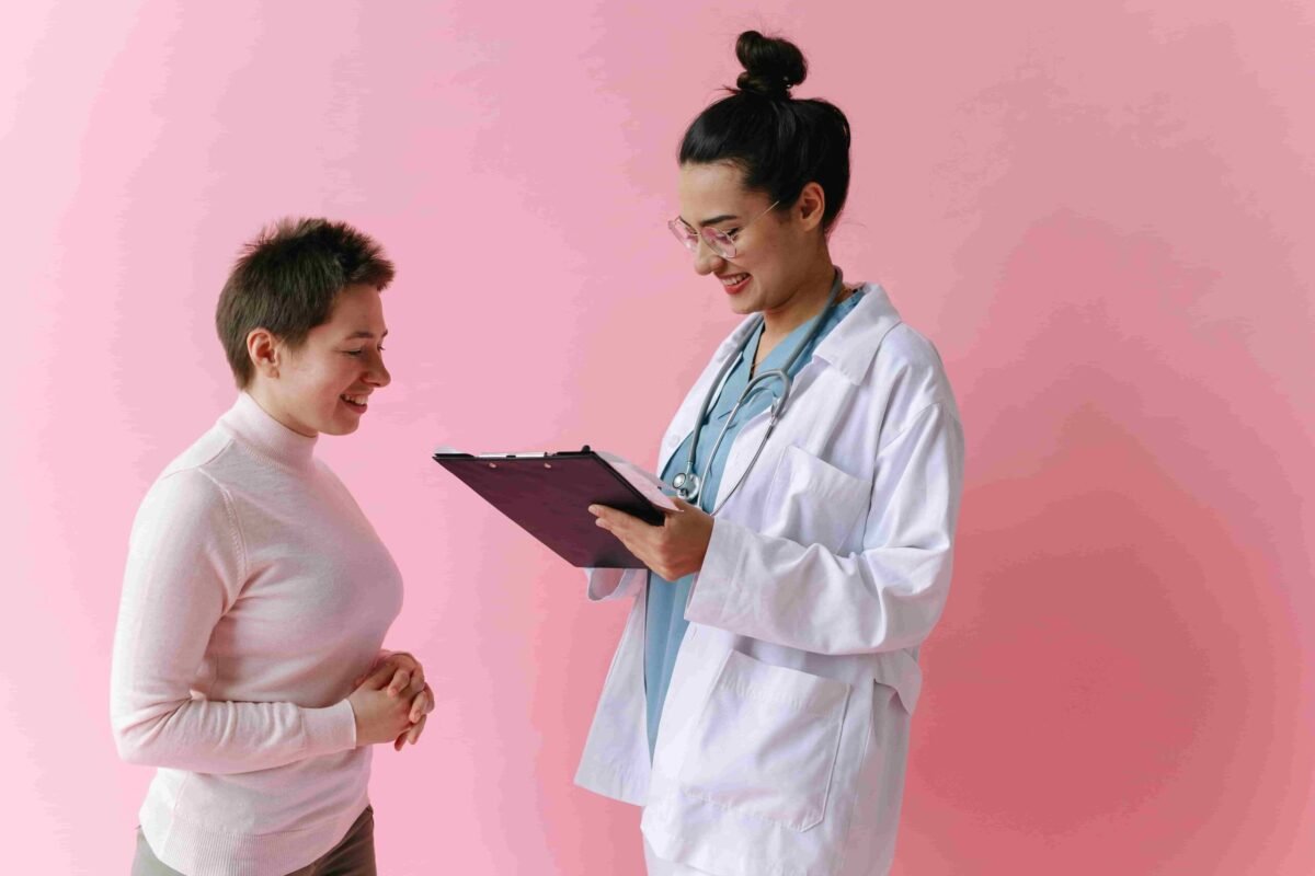 A doctor in a white coat holds a clipboard and speaks with a patient in a light pink sweater, both standing against a pink background. Find Direct Primary Care Providers in Las Vegas for personalized healthcare solutions.