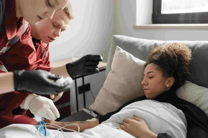 Two medical workers at an urgent care facility attend to a woman lying on a bed with an IV in her arm, closely monitoring her condition and providing care.