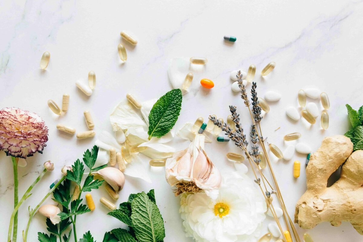 Assorted pills, capsules, herbs, flowers, garlic, and ginger root arranged on a white surface reflect the holistic approach of Integrative Medicine.