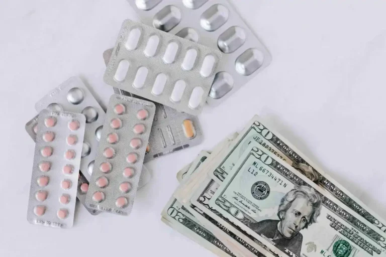 Several blister packs of medication are scattered next to a stack of U.S. twenty dollar bills on a white surface, highlighting potential lab savings.