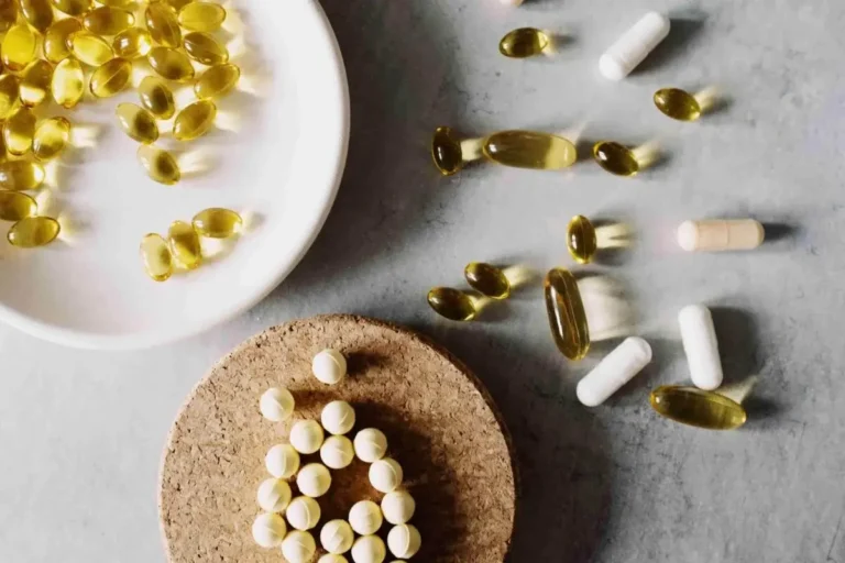 Assorted capsules and tablets, including yellow softgels and white pills—some inspired by stem cell products—are arranged on a white plate, a cork mat, and a gray surface.