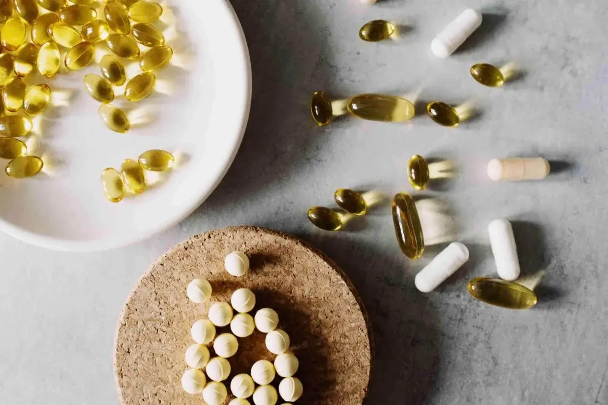 Assorted capsules and tablets, including yellow softgels and white pills—some inspired by stem cell products—are arranged on a white plate, a cork mat, and a gray surface.