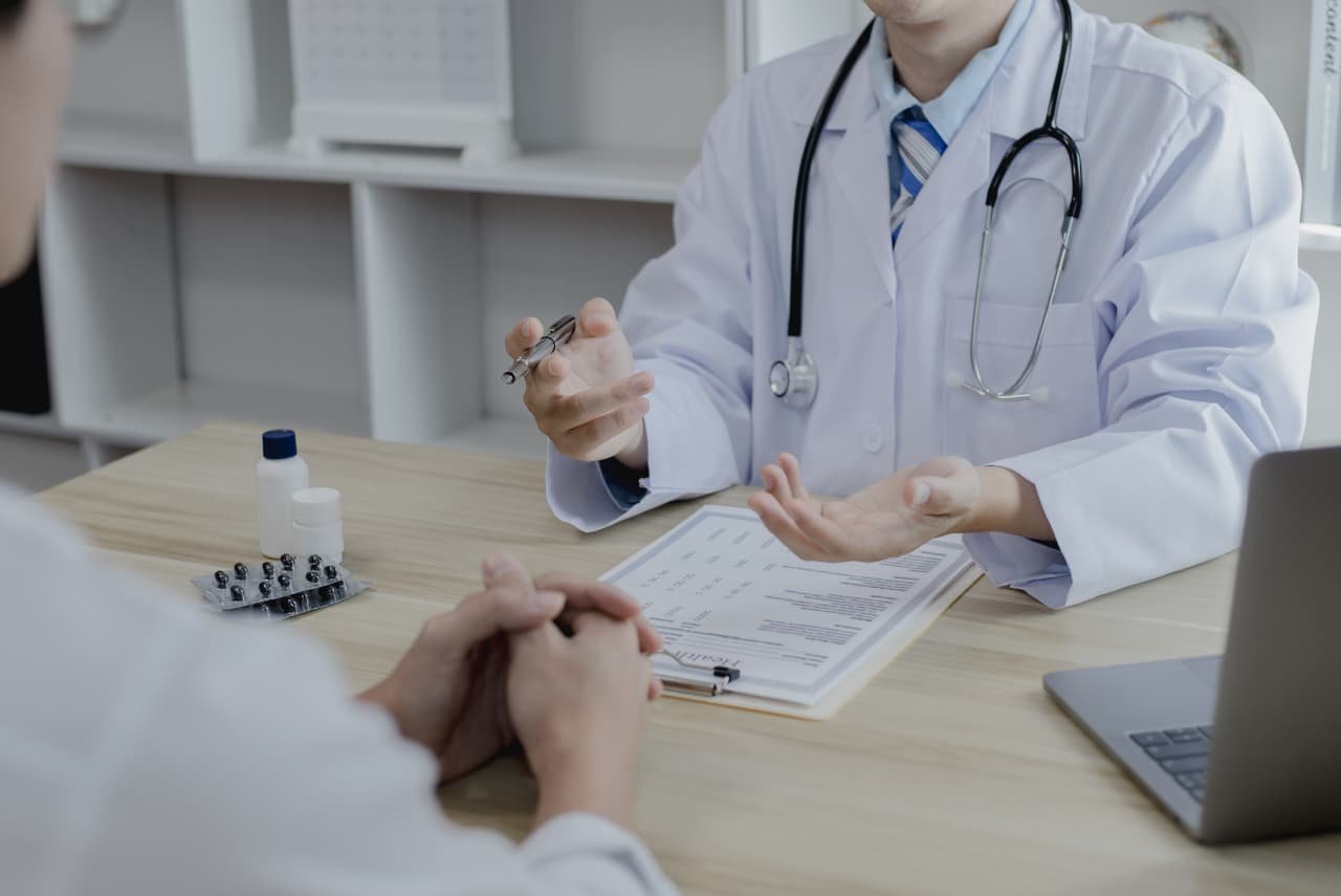 A doctor in a white coat discusses medical information with a patient across a desk, highlighting the benefits of Direct Primary Care, with a clipboard, medication, and a laptop on the table.