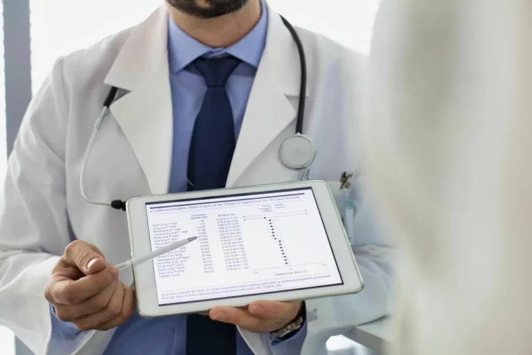 A doctor in a white coat and stethoscope holds a tablet displaying medical data, pointing at the screen while discussing Functional Medicine Las Vegas options with a patient.