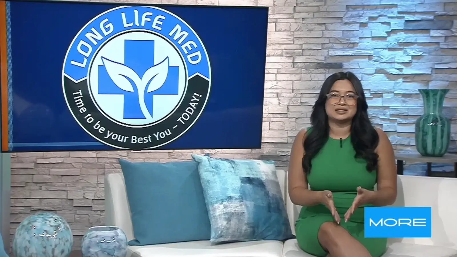 A woman in a green dress sits on a white couch beside blue pillows, with a “Long Life Med” logo displayed on a screen behind her during a Fox5 TV segment.